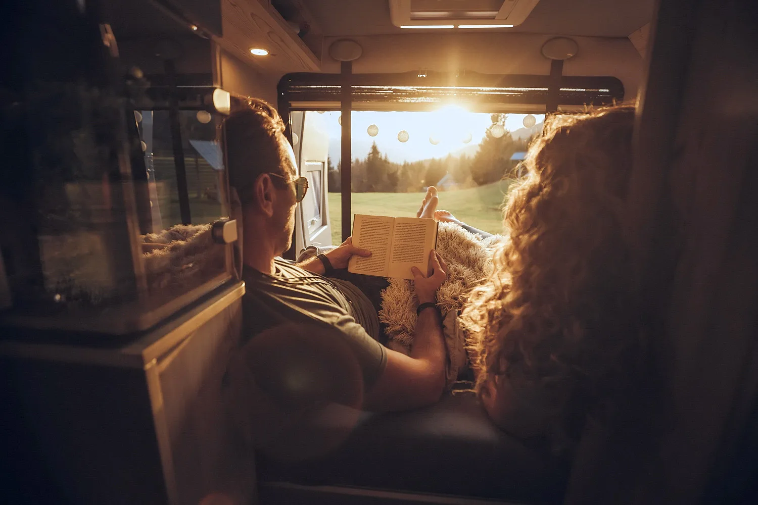 Couple relaxing inside a camper van with the man reading a book while the woman looks outside at a sunny countryside landscape.