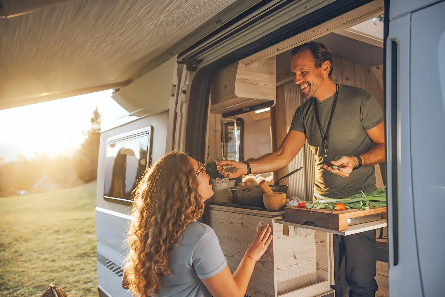 Man feeding woman by hand from the kitchen window of a camper van at sunset.