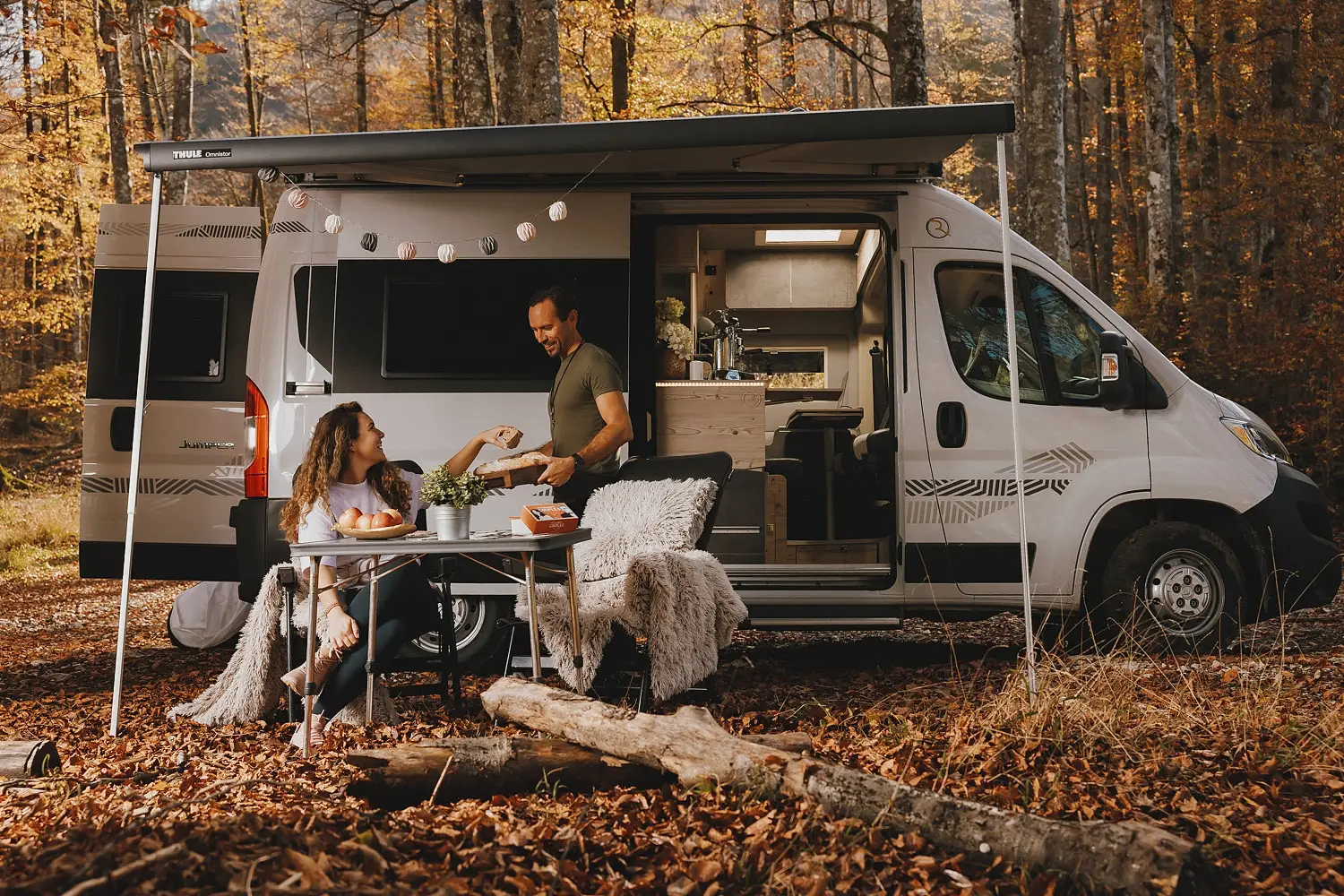 Couple enjoying a snack outside their camper van in a forest with autumn leaves.