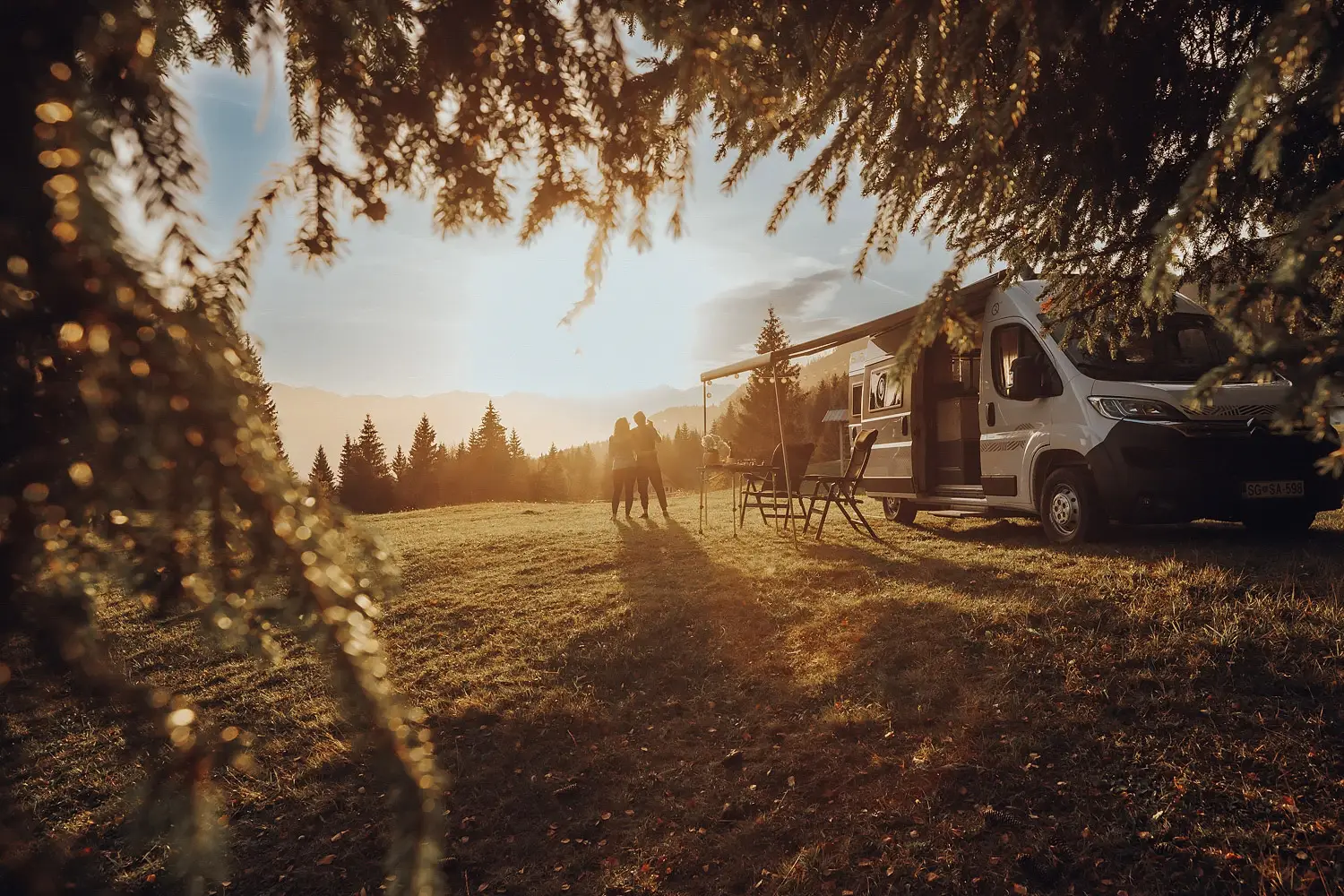 Two people embracing by a camper van with chairs and a table, framed by tree branches at sunset in a mountainous forest clearing.