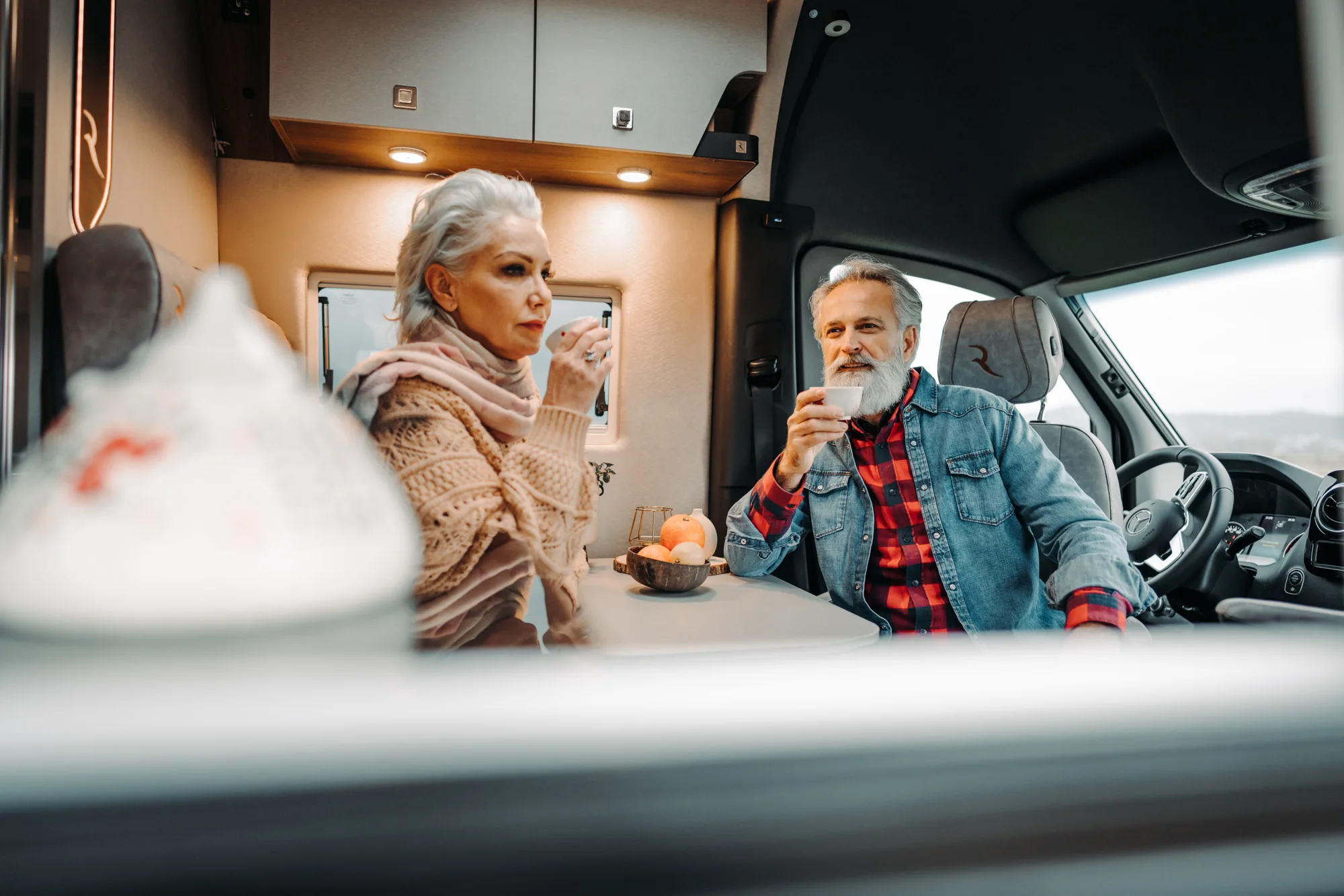 Older man and woman sitting inside a camper van drinking coffee at a table with a bowl of fruit.
