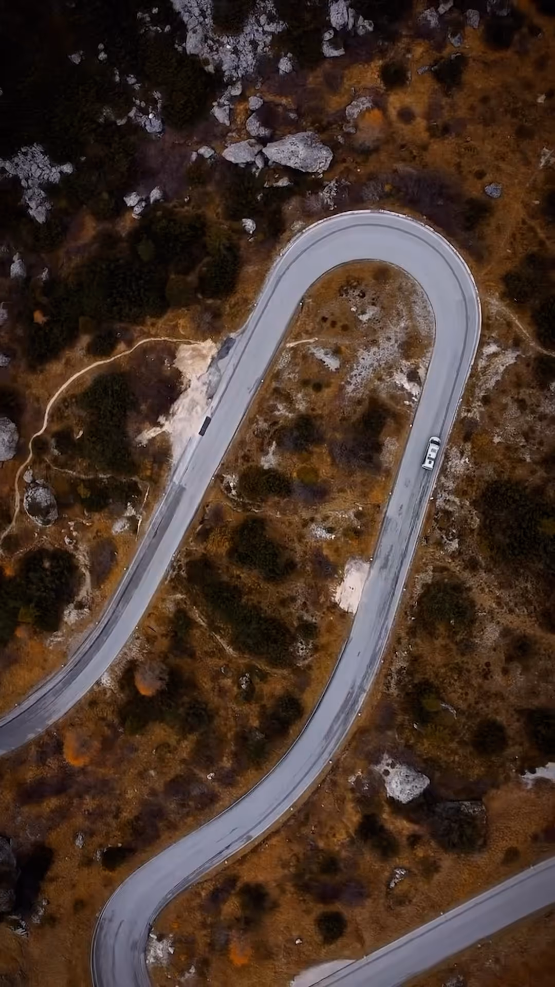 Aerial view of a winding mountain road surrounded by rocky and brown terrain with sparse vegetation and a white vehicle driving on the road.