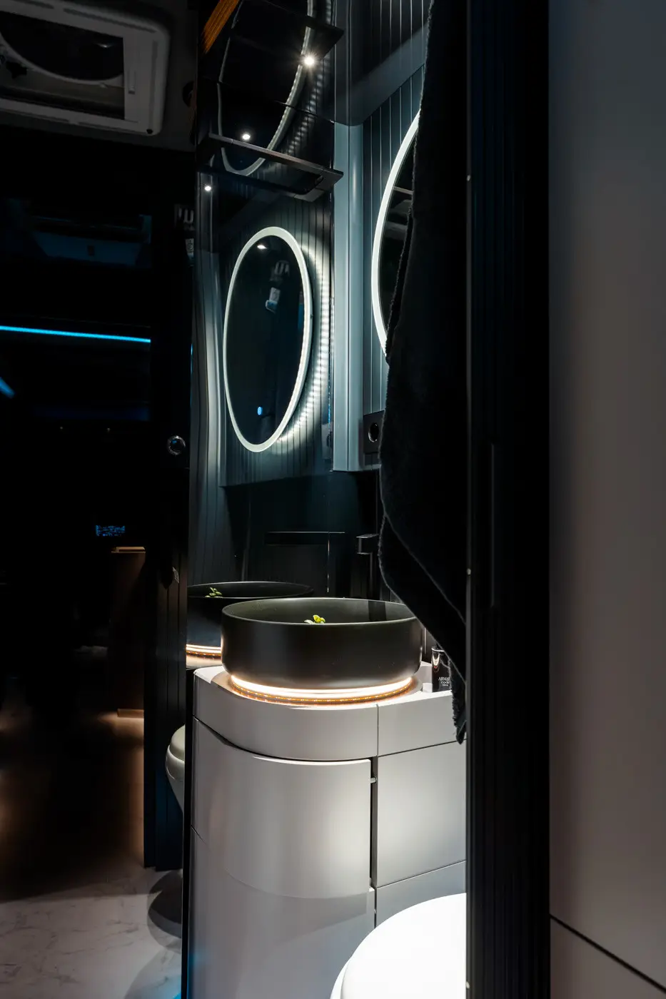 Bagno moderno con lavabo rotondo nero illuminato da strisce LED, specchio circolare retroilluminato e pannelli verticali scuri.