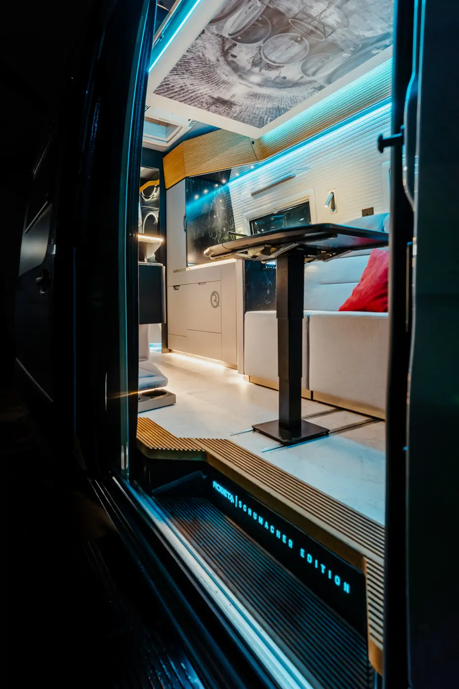 Vista interna di un moderno camper con tavolo da pranzo, panca bianca imbottita con cuscino rosso e illuminazione ambientale blu.