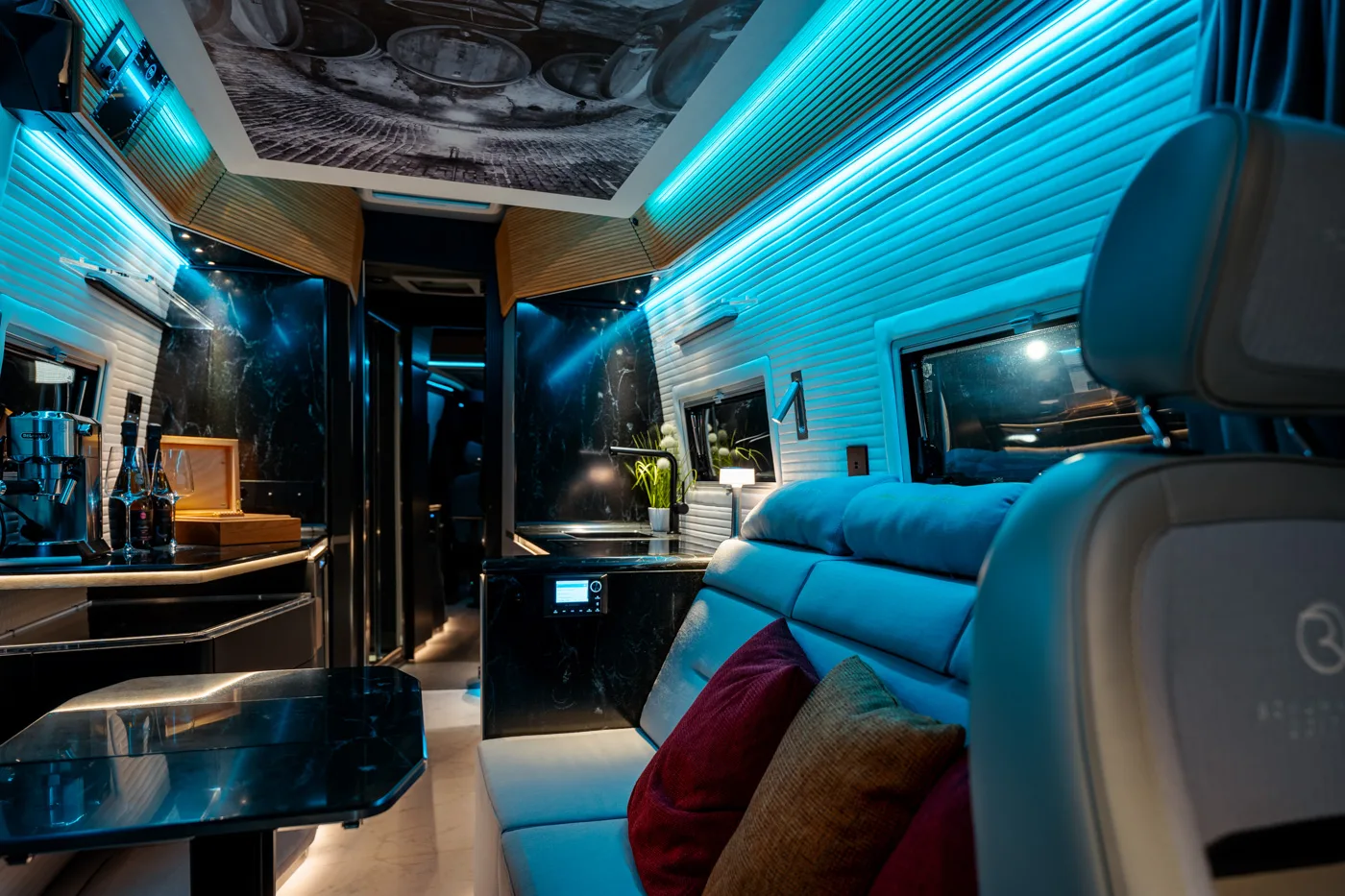 Lussuoso interno moderno di un camper con illuminazione a LED, superfici in marmo nero, sedute beige con cuscini colorati e un piccolo angolo cottura.