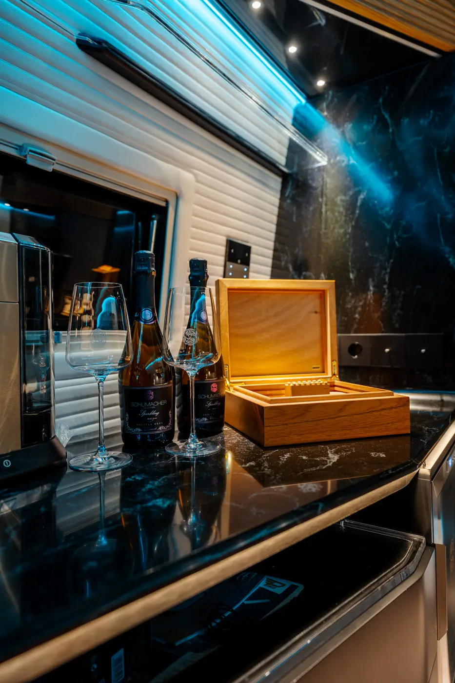 Due bottiglie di spumante Schumacher, due bicchieri vuoti e una cassetta di legno aperta su un piano di marmo nero con illuminazione blu.