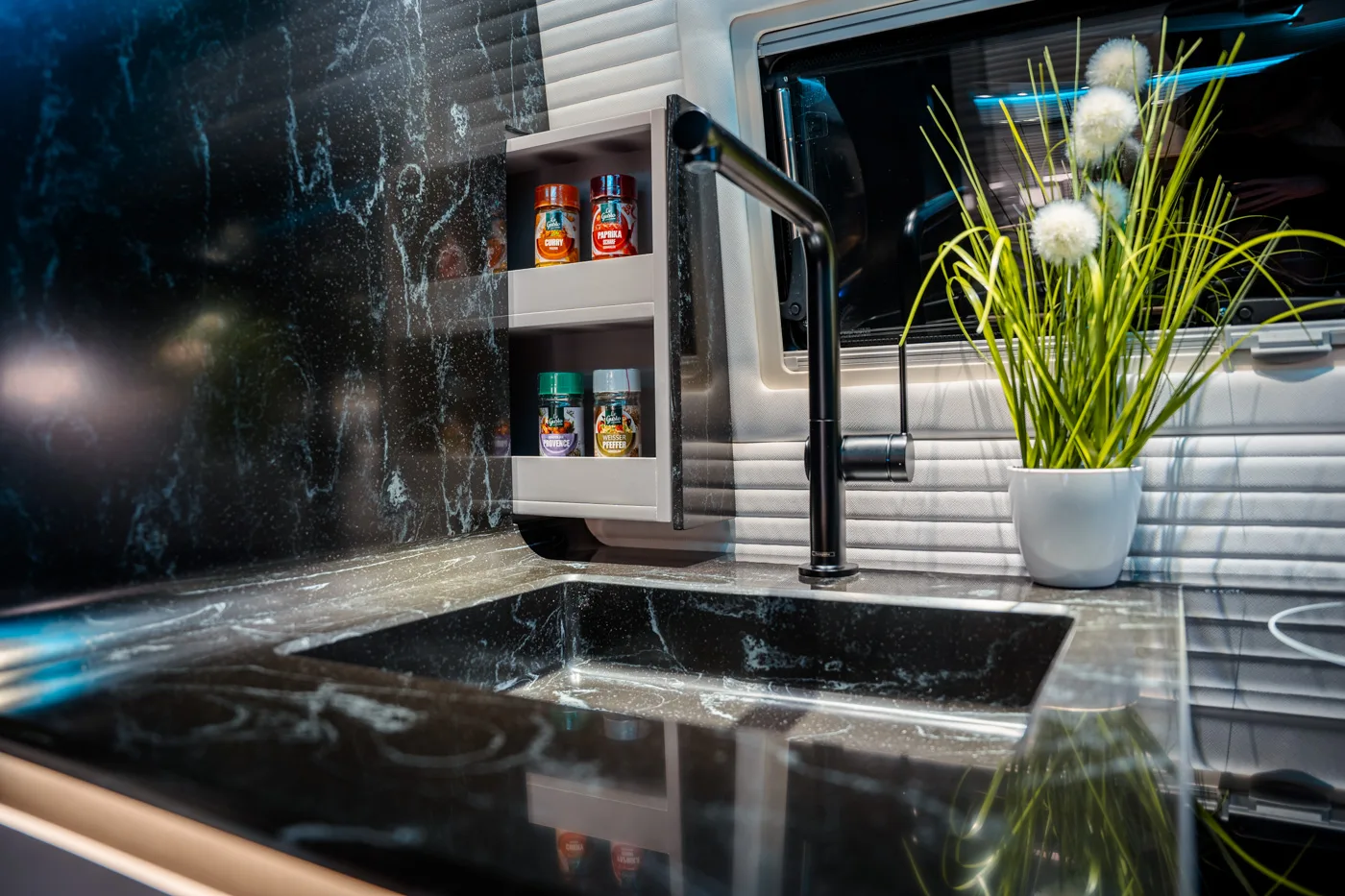Lavello da cucina moderno con piano in marmo nero, rubinetto nero, barattoli di spezie su una mensola bianca e un vaso bianco con erba verde decorativa e pon-pon bianchi.