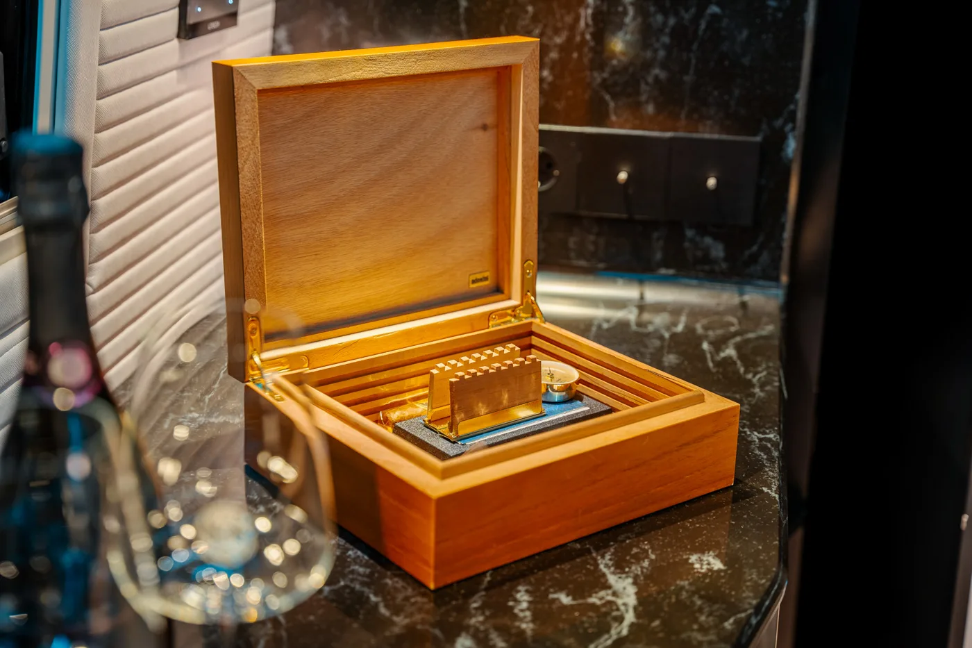 Scatola humidor in legno aperta su un piano di marmo con all'interno un sigaro e un igrometro, con una bottiglia di vino e un bicchiere sfocati in primo piano.