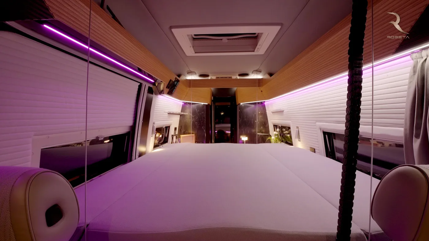 Interni moderni di un camper con letto spazioso, inserti in legno e illuminazione a strisce LED viola.