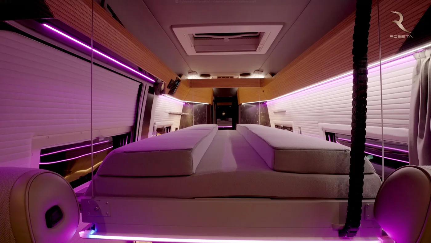 Interno moderno di un camper con letto rialzato incorniciato da luci LED viola e accenti in legno.