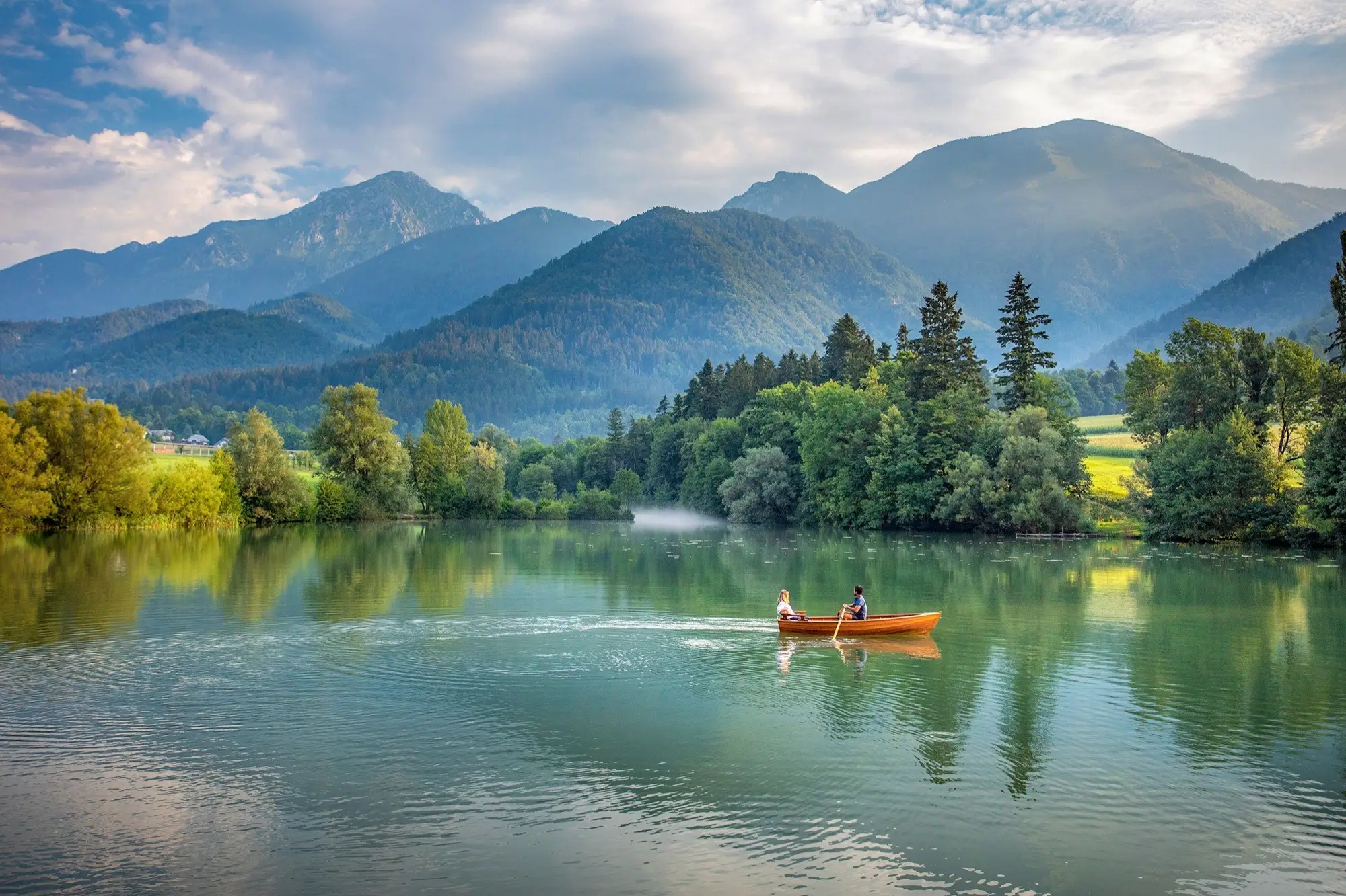 Two people rowing a wooden boat on a calm lake surrounded by lush green trees and mountains under a partly cloudy sky.