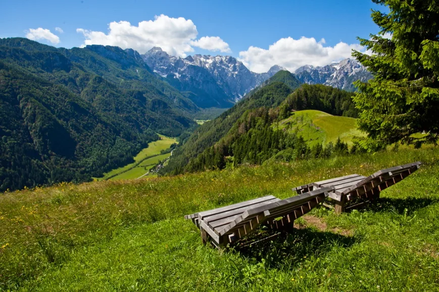 Two wooden lounge chairs on a grassy hillside overlooking green mountains and a partly cloudy blue sky.