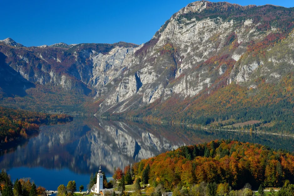 Tranquil lake reflecting rocky mountains and autumn foliage with a small church in the foreground.