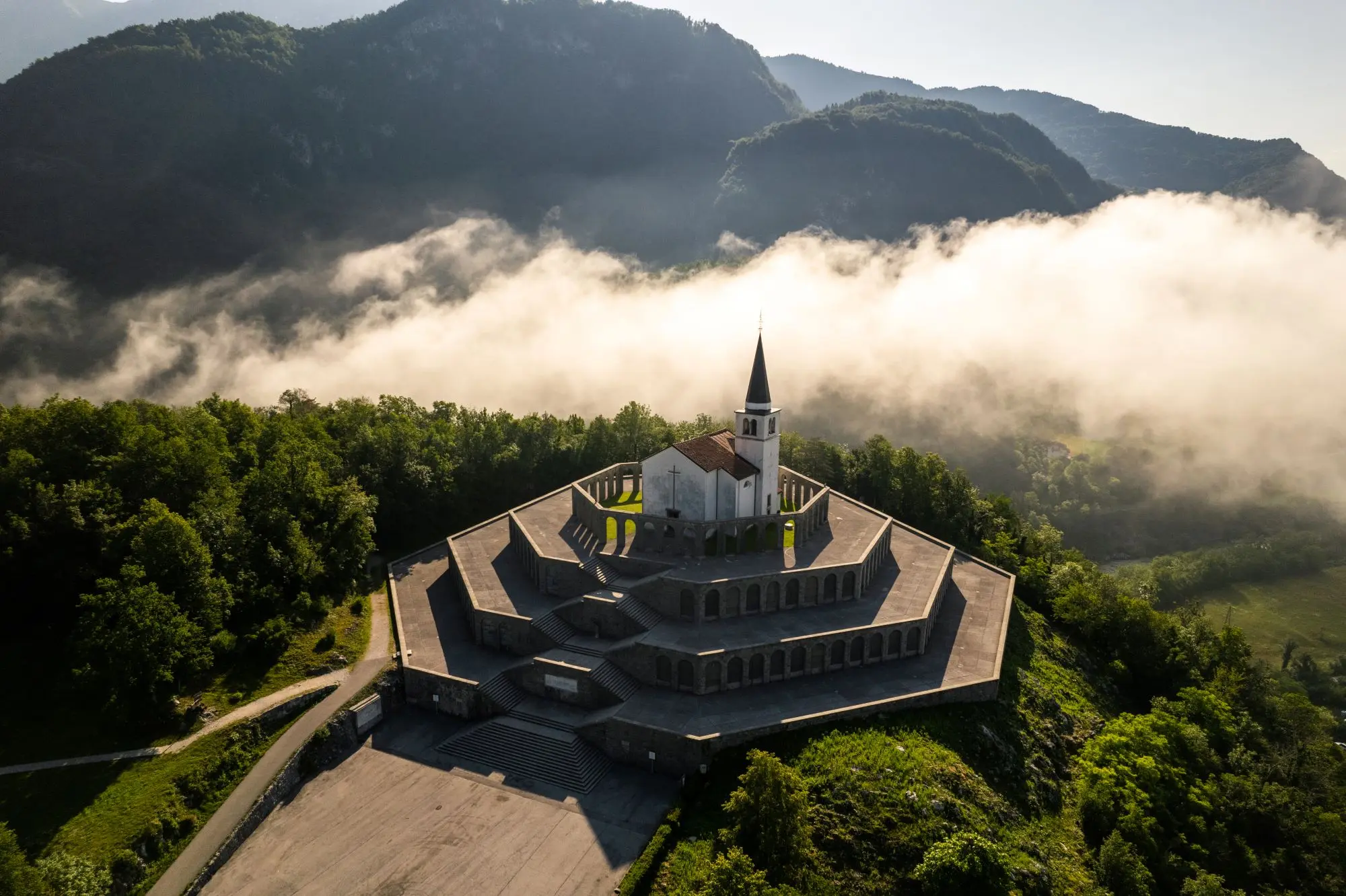 Drone view of a white church with a tall steeple atop a multi-tiered hexagonal stone platform surrounded by lush green trees and mountains with mist.