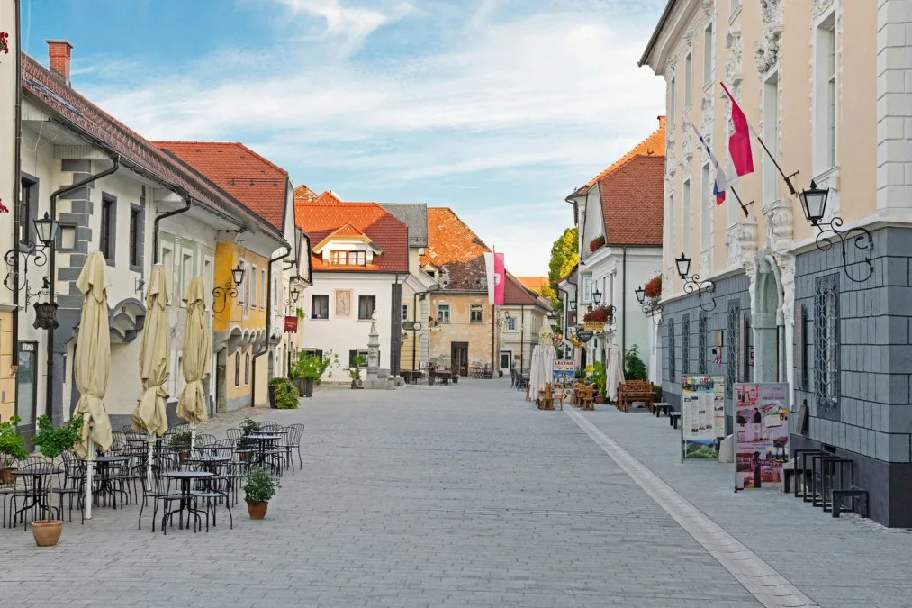 Calm pedestrian street with outdoor café tables and chairs, historic buildings with red roofs, and flags on a clear day.