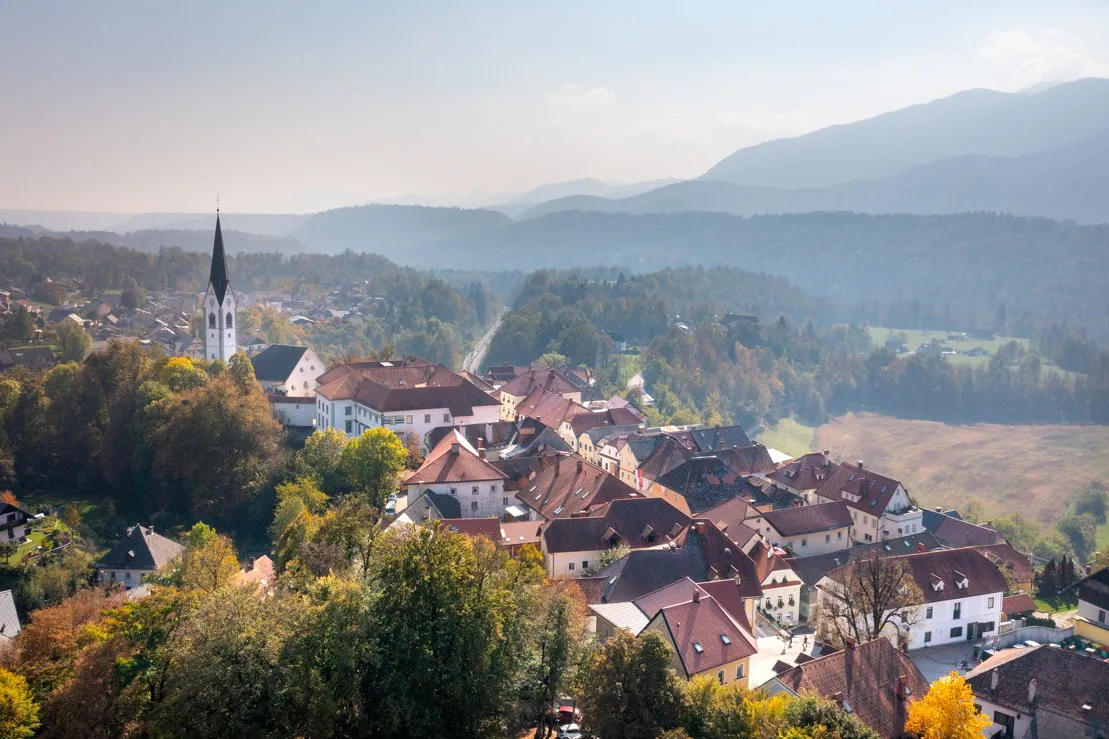 A quaint village with red-roofed houses and a tall church steeple surrounded by trees and hills under a clear sky.