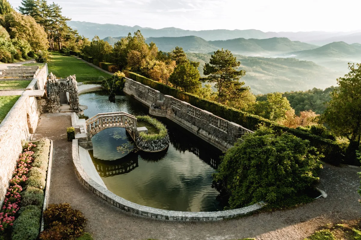 Stone bridge over a decorative pond in a terraced garden with trees and mountains in the background.