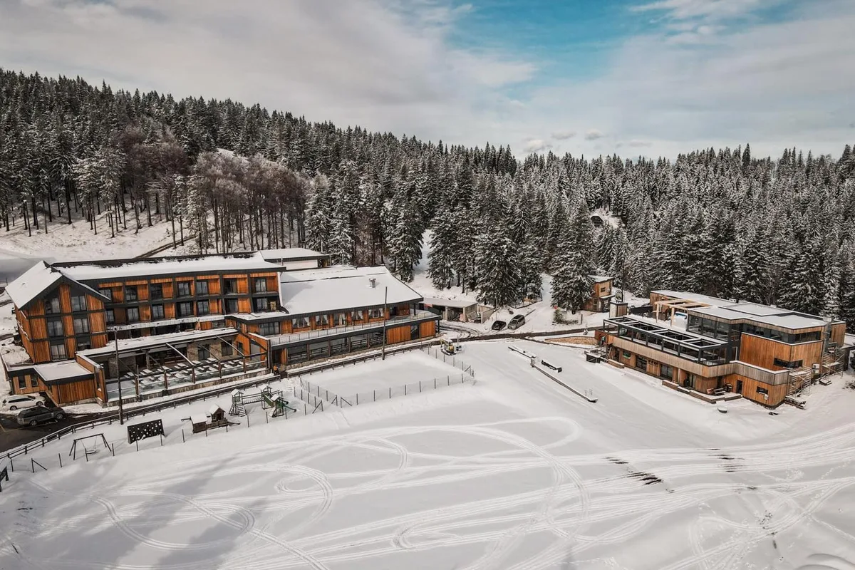 A wooden lodge surrounded by snow-covered ground and dense snow-laden pine trees under a partly cloudy sky.