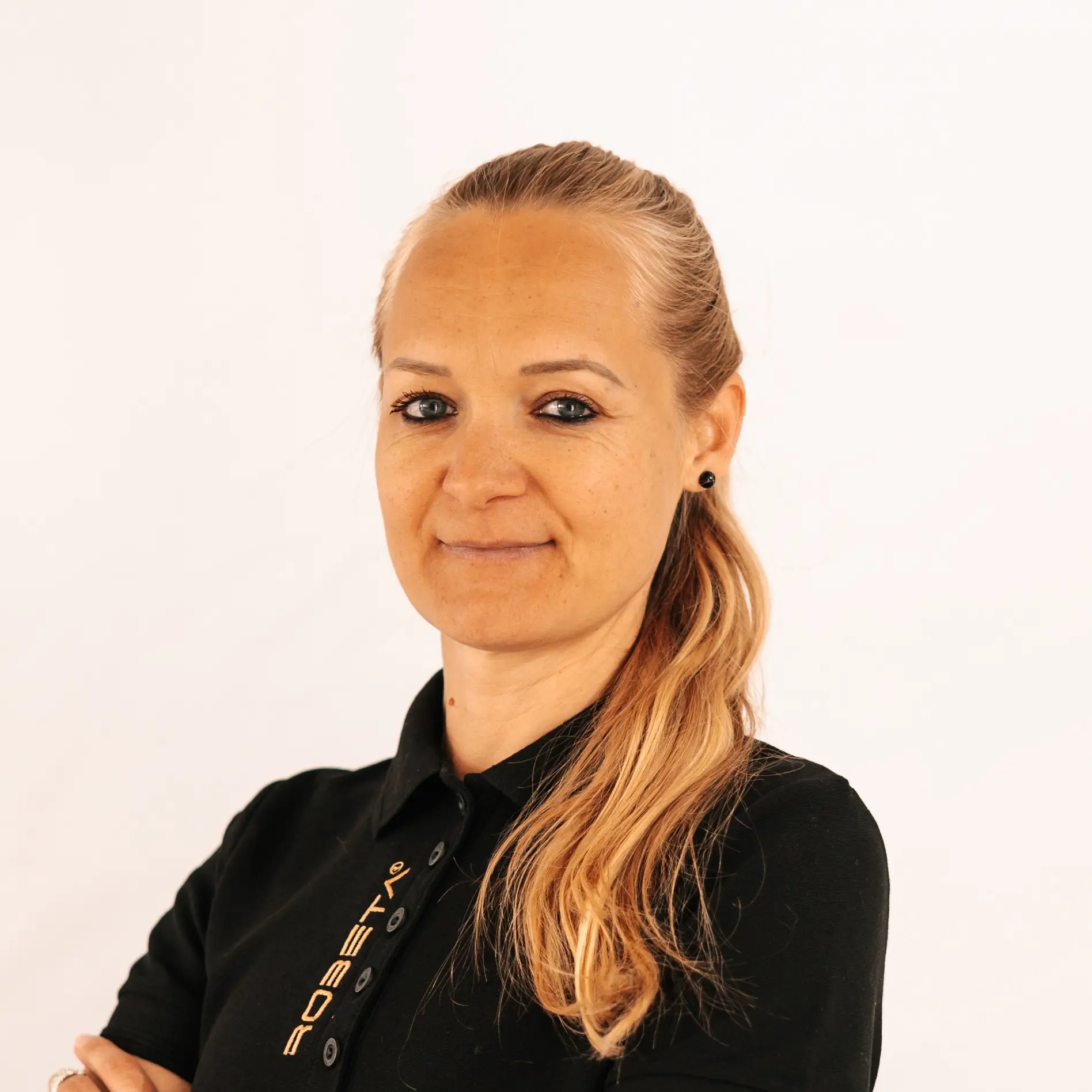 Smiling woman with long blonde hair in a black polo shirt with crossed arms against a plain white background.
